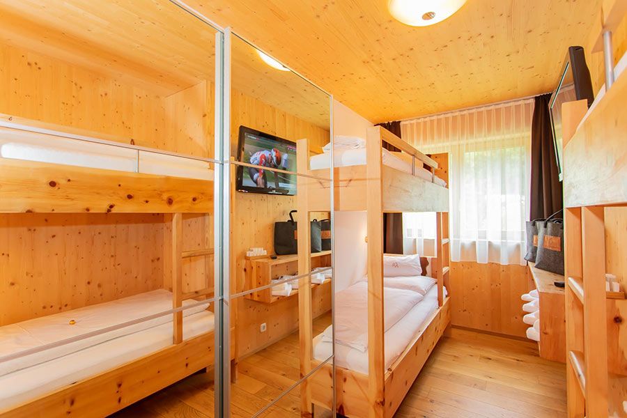 Kleines Zimmer mit zwei Etagenbetten aus hellem Holz, Spiegelkleiderschrank, Fernseher an der Wand und Fenster mit Gardinen