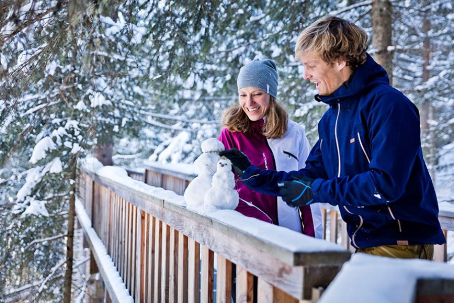 Zwei Personen in Winterkleidung bauen auf einem schneebedeckten Holzgeländer kleine Schneemänner
