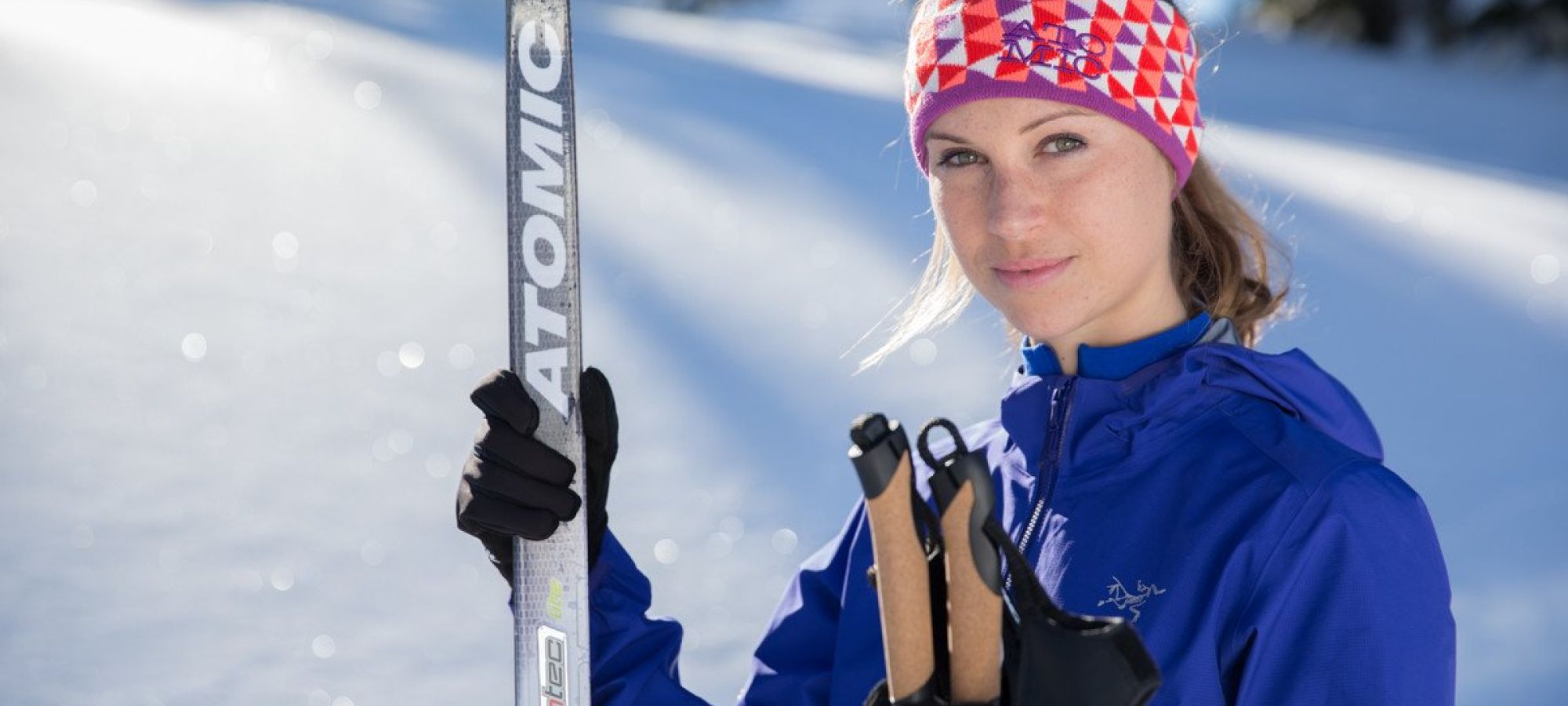 Person in blauer Skijacke und buntem Stirnband hält einen Skistock und einen Ski mit der Aufschrift 'ATOMIC' im Schnee