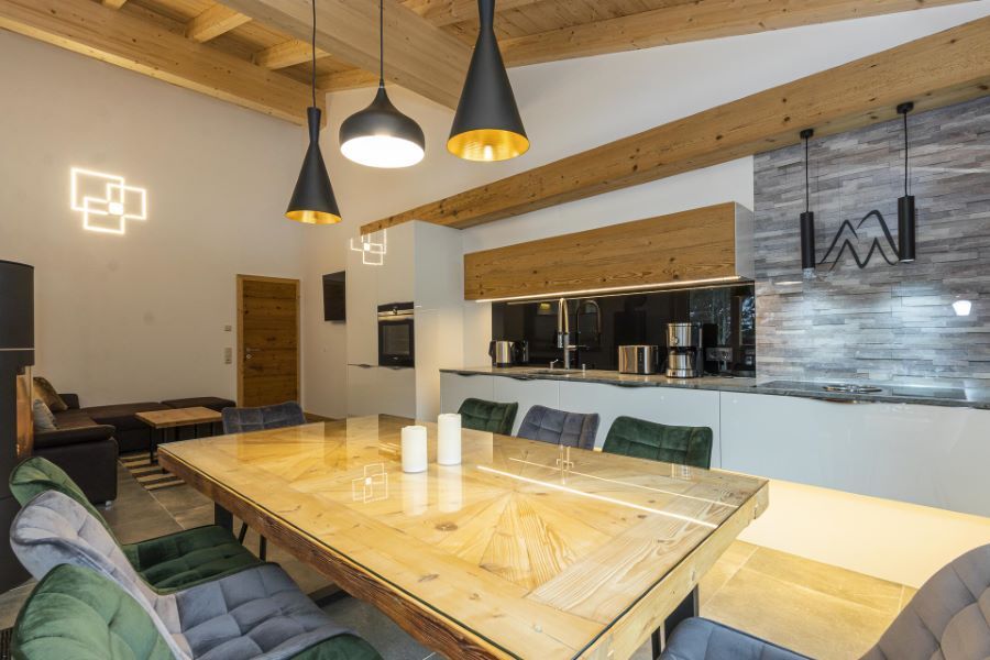 Moderner Essbereich mit großem Holztisch, gepolsterten Stühlen, offener Küche mit weißen Fronten und Holzdecke mit sichtbaren Balken