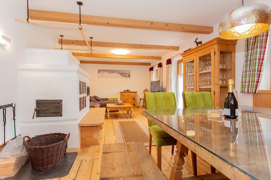 Dining area with glass-top table and green chairs next to a wooden cabinet filled with glassware and a white tiled stove with a basket on the floor
