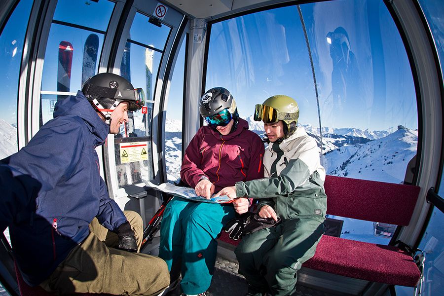 Drei Personen mit Skihelm und Skibekleidung sitzen in einer Gondelbahn und betrachten eine Karte vor verschneiter Berglandschaft