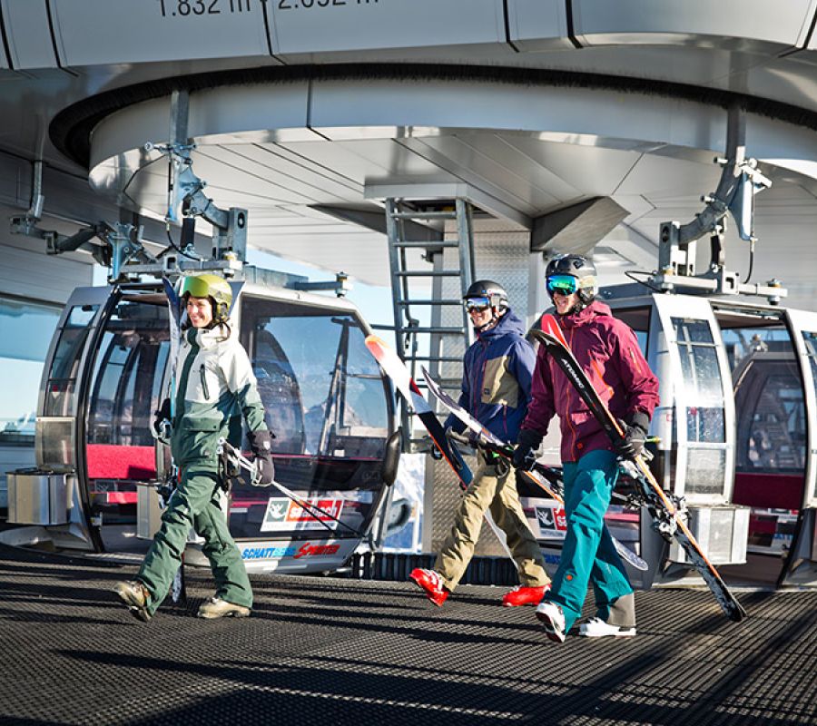 Drei Personen in Skibekleidung mit Skiern gehen vor Gondelbahnstation auf schwarzem Gitterboden