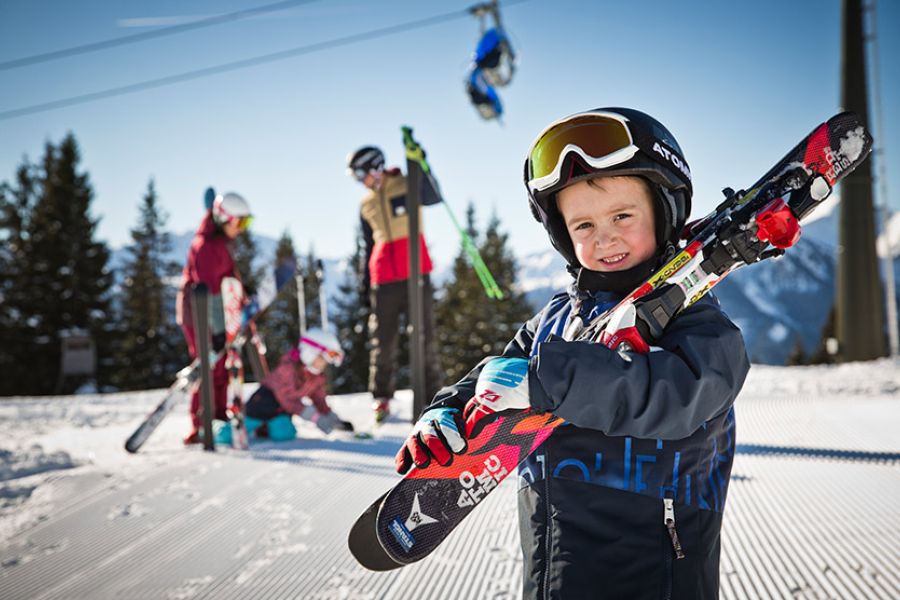 Kind mit Skihelm und Skibrille trägt Skier auf der Schulter auf einer schneebedeckten Skipiste mit weiteren Personen im Hintergrund