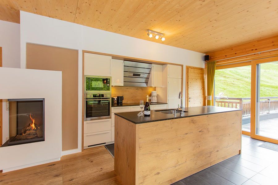 Moderne Küche mit Kochinsel aus Holz, eingebautem Ofen, Kamin mit Feuer und großer Glas-Schiebetür zum Balkon mit Blick auf eine grüne Wiese